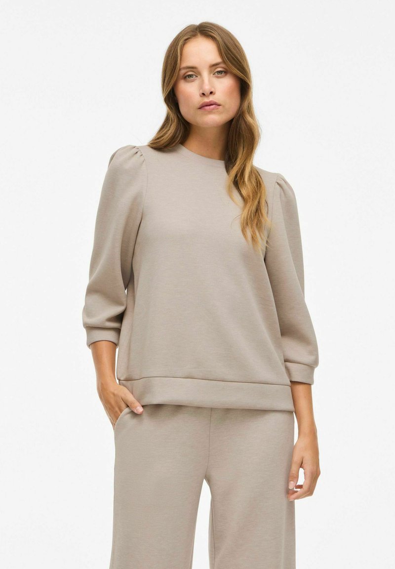 Felpa beige con scollatura rotonda, maniche a tre quarti a sbuffo e vestibilità rilassata. Realizzata in tessuto morbido e liscio con orlo a strati.