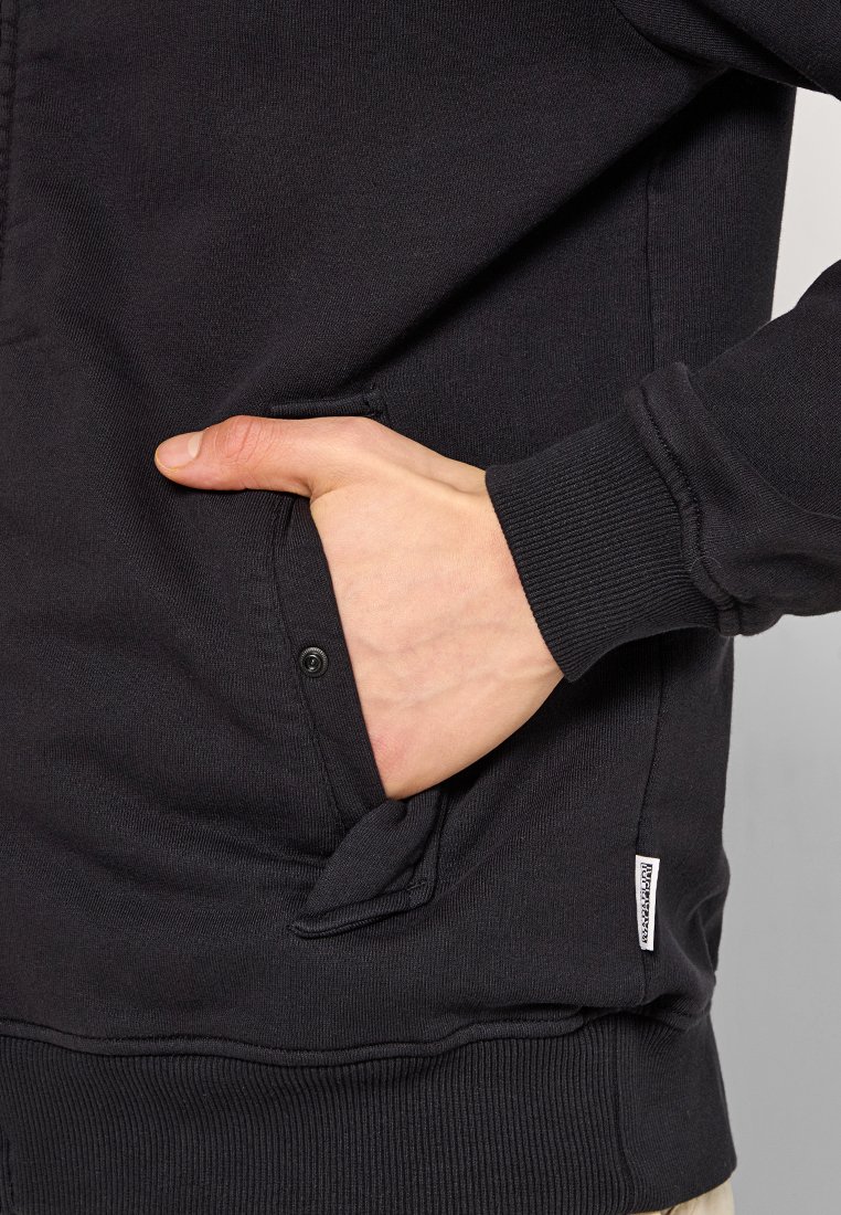 Hand in der Vorderentasche eines schwarzen Hoodies mit geripptem Bündchen, Knopfdetails und kleinem weißen Markenschild am Saum.