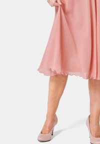 Rosa Chiffonkleid mit einem fließenden, geschichteten Rock und durchscheinendem Saum; kombiniert mit hellrosa Wildledersandalen. Fokus auf Textur und Farbkontrast.