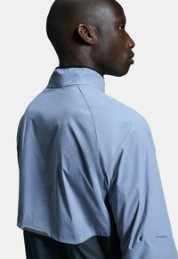 Hellblaues Jackett mit hohem Kragen und tailliertem Design, mit glatter Textur, Nahtdetails und einem Logo auf dem Ärmel.