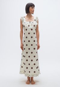 Γυναίκα με κοντά σκούρα μαλλιά που φοράει αμάνικο maxi φόρεμα κρεμ με μεγάλα καφέ πουά, με βολάν στο τετράγωνο ντεκολτέ και στις άκρες, όρθια και ξυπόλυτη.