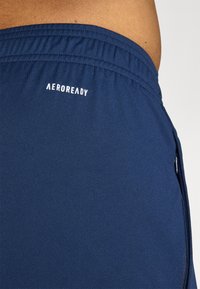 Mørkeblå træningsshorts med elastisk talje. Har "AEROREADY"-logo i hvid og en sidelomme med lynlås til opbevaring. Glat tekstur.