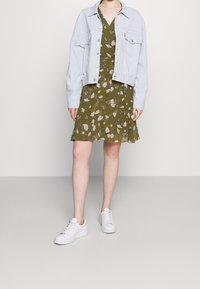 Světle modrá džínová bunda přes zelené květinové šaty s volánkovým lemem. Bílá tenisky dokončují outfit. Měkká tkanina a volný střih.