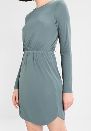 Woman wearing a long-sleeve, knee-length, sage green dress with gathered waist and rounded hem, standing against a plain background.