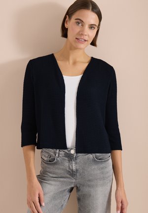 Femme aux cheveux courts et bruns portant un cardigan noir texturé ouvert sur le devant, un haut blanc et un jean gris clair sur un fond beige.