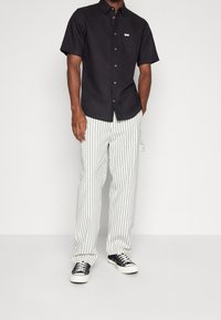 Chemise noire à manches courtes avec une poche avant, associée à un pantalon blanc à rayures. Les baskets ont une tige noire et une semelle blanche.