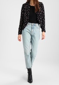 Blazer noir à motif floral, haut noir, jean taille haute bleu clair avec ourlet effiloché. Le mannequin porte des bottines noires à talons. Fleurs de différentes couleurs sur la veste.