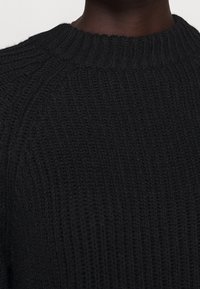 Schwarzer Strickpullover mit strukturierter Rippoptik und klassischem Rundhalsausschnitt. Der Stoff wirkt dick und warm, ideal für kältere Wetterbedingungen.