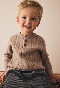 Tan cable-knit sweater with brown buttons, paired with gray corduroy pants. Child is seated, smiling, with short, tousled blonde hair.