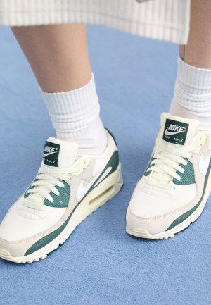 Stopy noszące białe buty Nike Air Max z zielonymi akcentami i białe prążkowane skarpetki stojące na niebieskim dywanie.