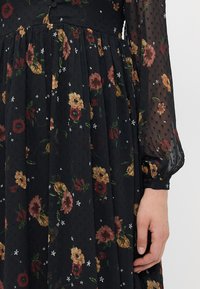Svart blomstret kjole med gjennomsiktige lange ermer. Har fargerike blomster mønstre i rødt, gult og hvitt, med et strukturert stoff og en innsnevret midje.