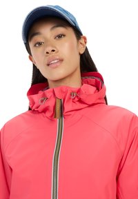 Bright pink waterproof jacket with a high collar and drawstrings, paired with a denim cap. Features a visible zipper and stitching details.