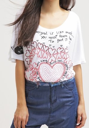 Frau trägt ein weißes Hemd mit rotem Herz und Schriftzügen, kombiniert mit blauen High-Waist-Jeans, steht mit entspannten Händen seitlich am Körper.