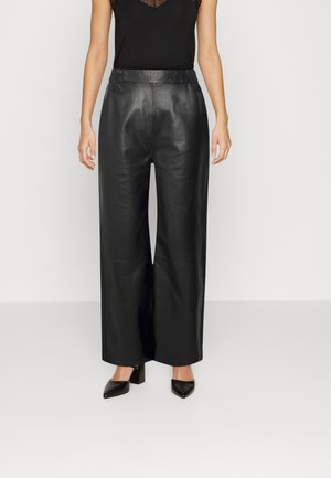 Femme portant un haut noir sans manches avec un détail en dentelle et un pantalon en cuir noir à jambes larges, debout devant un fond clair uni.