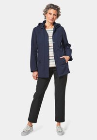Navy wasserfeste Jacke mit Kapuze, Reißverschluss und Knopfverschlüssen, zwei Fronttaschen, kombiniert mit einem gestreiften Hemd und schwarzen Hosen, getragen mit Sneakers.