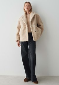 Veste en shearling beige en peluche avec un large col, deux poches avant et des boutons argentés, portée avec un pull léger et un jean noir à longueur de cheville.