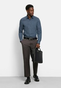 Ein dunkelblaues Hemd mit geometrischem Muster, kombiniert mit dunklen Hosen und schwarzen Schuhen, während er eine schwarze Ledertasche hält.