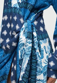 Μπλε ύφασμα με σχέδια παϊλεϋ και λουλουδιών, με δεμένη ζώνη στη μέση και αέρινες μανσέτες. Τα υλικά φαίνονται ελαφριά και λεία.