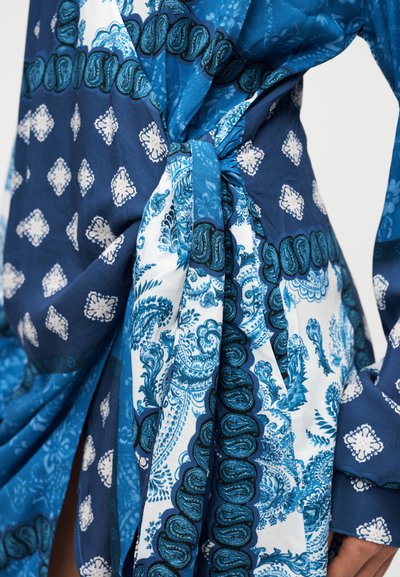 Μπλε ύφασμα με σχέδια παϊλεϋ και λουλουδιών, με δεμένη ζώνη στη μέση και αέρινες μανσέτες. Τα υλικά φαίνονται ελαφριά και λεία.