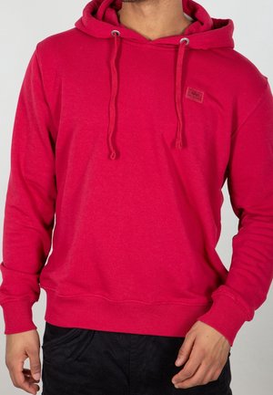 Mężczyzna w jaskrawoczerwonej bluzie z kapturem ze ściągaczami i małą naszywką na piersi, w czarnych spodniach, stojący z rękami blisko kieszeni.