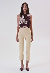 Top senza maniche in seta marrone scuro con stampa floreale; pantaloni gialli aderenti con dettagli di bottoni all'orlo; abbinati a ballerine marroni scuri.