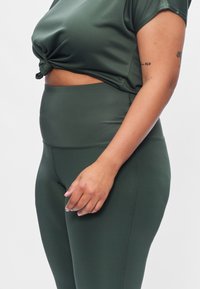 Donkergroene sporttop met een geknoopt ontwerp in de taille, gecombineerd met hooggetailleerde donkergroene leggings van gladde, rekbare stof.