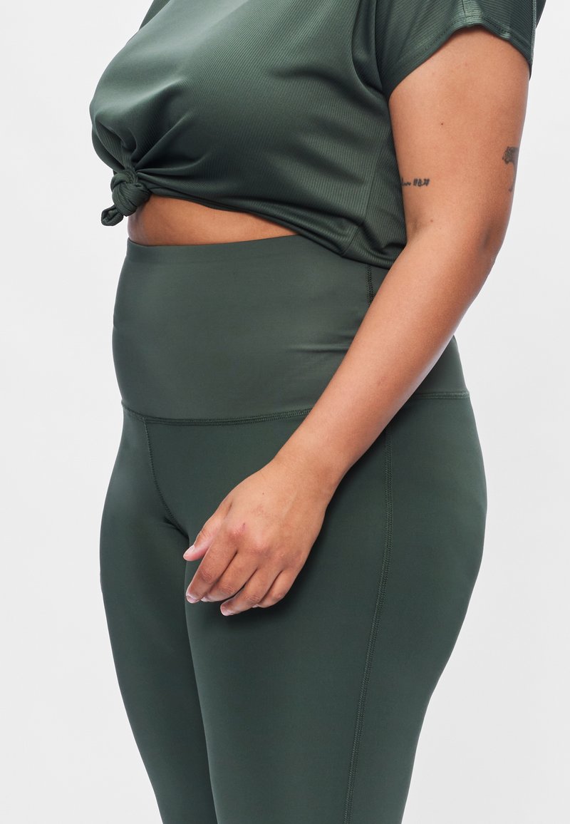 Donkergroene sporttop met een geknoopt ontwerp in de taille, gecombineerd met hooggetailleerde donkergroene leggings van gladde, rekbare stof.