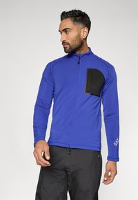 Plava sportska zip-up jakna od glatkog materijala, s crnim džepom na grudima i kontrastnim logom na rukavu.