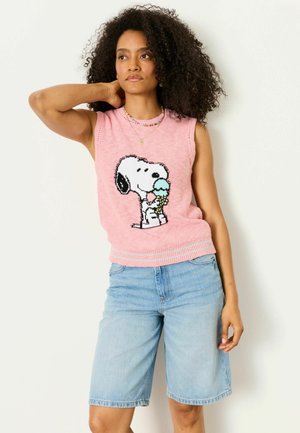Frau mit lockigem Haar, die einen rosa ärmellosen Pullover mit Snoopy, der Eis isst, und helle, knieumspielende Jeansshorts trägt.