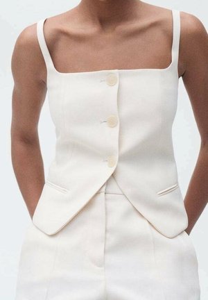 Femme portant un gilet blanc sans manches à trois boutons et un pantalon taille haute assorti, mains dans les poches, fond neutre.