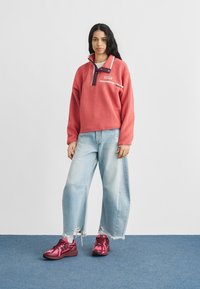 Γυναίκα στέκεται φορώντας ένα κορυφαίο από φλίς κοραλλί, ανοιχτό μπλε τζιν με φαρδιά πόδια και σκισίματα, και ροζ αθλητικά παπούτσια, πάνω σε μπλε χαλί.