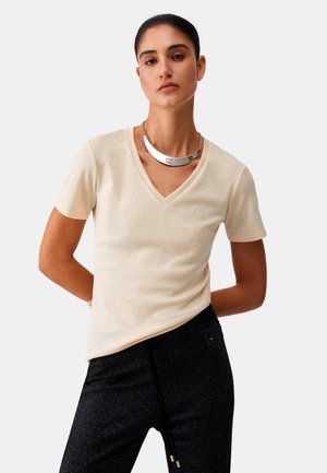 Donna con capelli scuri, indossa una maglietta beige a maniche corte con scollo a V e pantaloni neri, con una collana a catena d'argento, in piedi con le mani dietro la schiena.