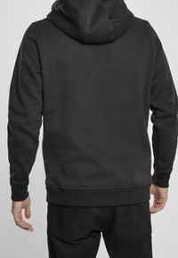 Schwarzer Hoodie mit langen Ärmeln und Kapuze, aus einem glatten Stoff gefertigt. Verfügt über gerippte Bündchen und Saum. Kein sichtbares Logo. Wird mit dunkler Hose getragen.