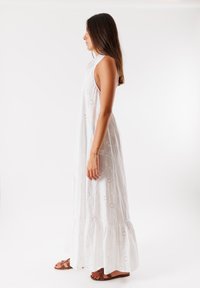 Abito lungo in cotone bianco con ricami ad occhio di piuma floreali, orlo a strati, design senza maniche e vestibilità расслабленные. Indossato con sandali marroni.