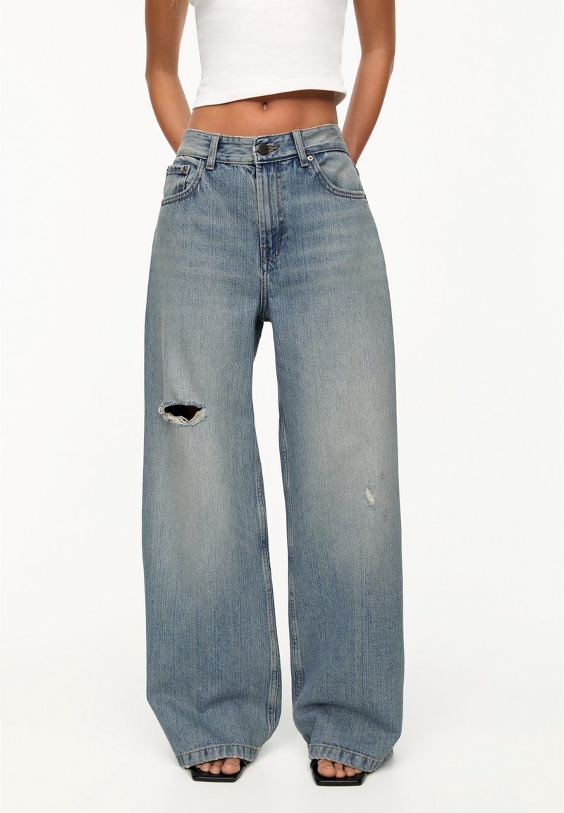 Femme portant un jean taille haute bleu clair à jambes larges avec un trou au genou et une déchirure effilochée à la cuisse, associé à un débardeur court blanc.