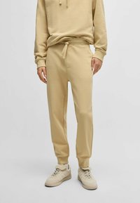 Beige katoenen joggers in een katoenmix met een elastische tailleband en een trekkoord, voorzien van zijzakken en gekraagde enkels. Gekleed met beige sneakers.