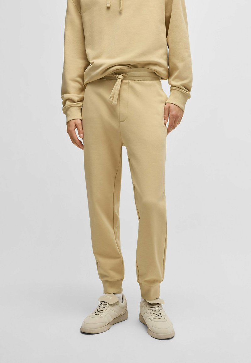Beige katoenen joggers in een katoenmix met een elastische tailleband en een trekkoord, voorzien van zijzakken en gekraagde enkels. Gekleed met beige sneakers.