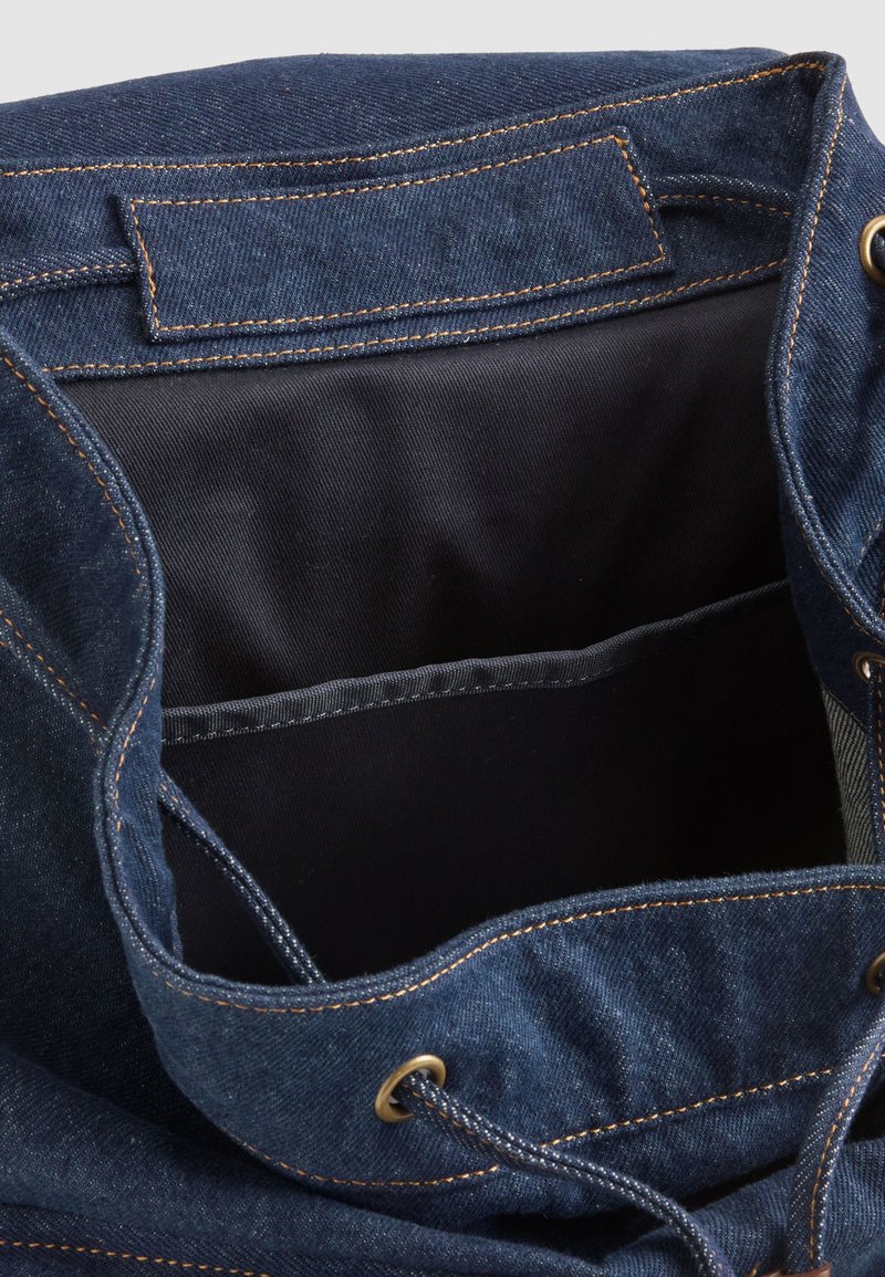 Interno della borsa a secchiello in denim, blu scuro con cuciture arancioni a contrasto, presenta una fodera interna nera e una tasca aggiuntiva per riporre gli oggetti.