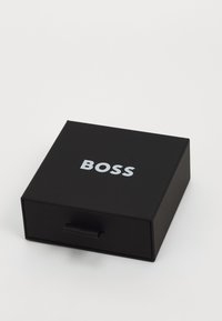 Svart boks med en jevn tekstur, med en glidende skuff og sølvfarget "BOSS"-logo på toppen. Kompakt rektangulær form.