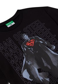 Černé bavlněné tričko s designem Superman v šedé barvě a červeným emblémem. Text zní "Muži z oceli" v opakujícím se vzoru.