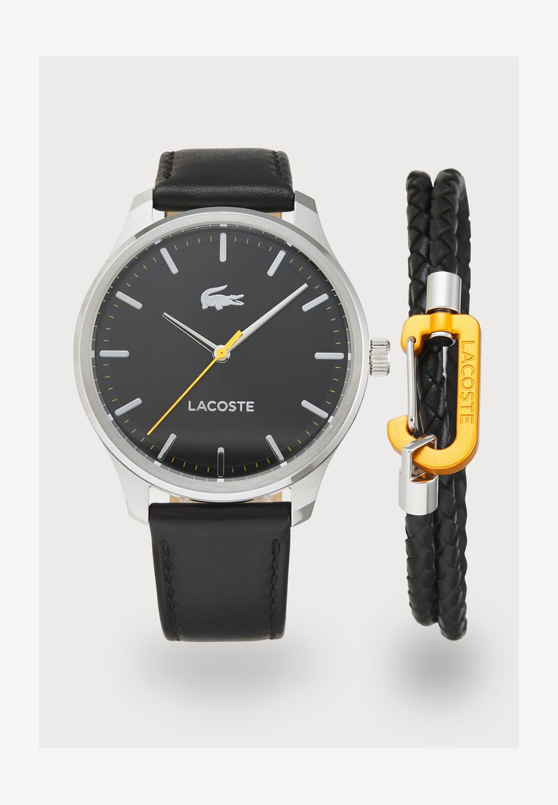 Relógio com pulseira de couro negro, caixa prateada, mostrador preto, ponteiros prateados e detalhes em amarelo. Inclui uma pulseira trançada preta com fecho laranja.