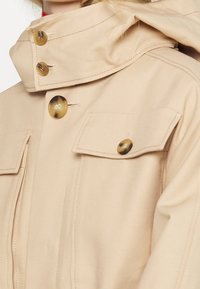Veste beige avec de gros boutons en écaille, poches à rabat boutonnées sur la poitrine, et un col montant qui masque partiellement le cou.