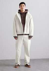 Chaqueta impermeable gris claro, sudadera marrón y pantalones crema; zapatos casuales con acentos marrones y beige; tejidos suaves y corte relajado.