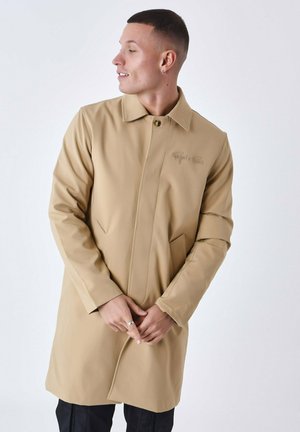 Chaqueta beige de manga larga con cuello de solapa, cierre de botones, tejido texturizado, bolsillos laterales y un sutil detalle de logo en el pecho.