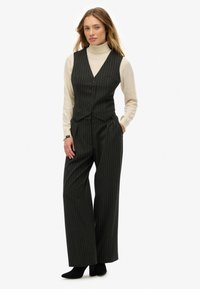 Mujer que lleva un chaleco de rayas negras y pantalones de pierna ancha con un suéter de cuello alto crema y botas de tacón negras, de pie con las manos en los bolsillos.
