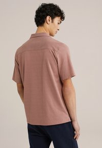 Polo a maniche corte di colore ruggine realizzato in un tessuto morbido con una texture liscia, caratterizzato da un colletto classico e dettaglio della cucitura sul retro.