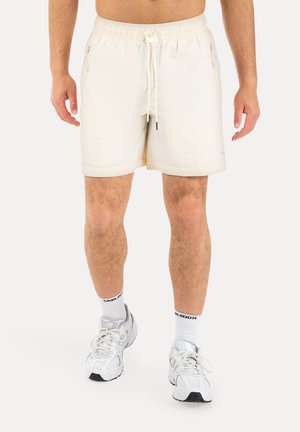 Mann steht und trägt cremefarbene Shorts mit Kordelzug, weiße Socken mit Text und weiße Sportschuhe auf weißem Hintergrund.