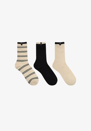 Trois chaussettes mi-mollet présentées côte à côte : beige avec des rayures noires, noir uni, et beige uni, chacune avec une petite étiquette noire en haut.