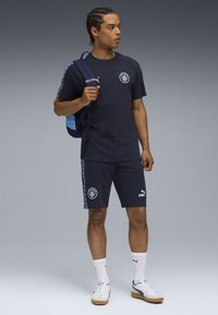 Marine atletik outfit med en kortærmet skjorte og matchende shorts. Har logo-detaljer, lyseblå accenter og en tekstureret rygsæk.
