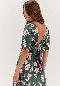 Zelené kvetinové šaty s krátkymi rukávmi, nízkym výstrihom na chrbte a viazaním v páse. Tkanina má hladkú, ľahkú štruktúru.